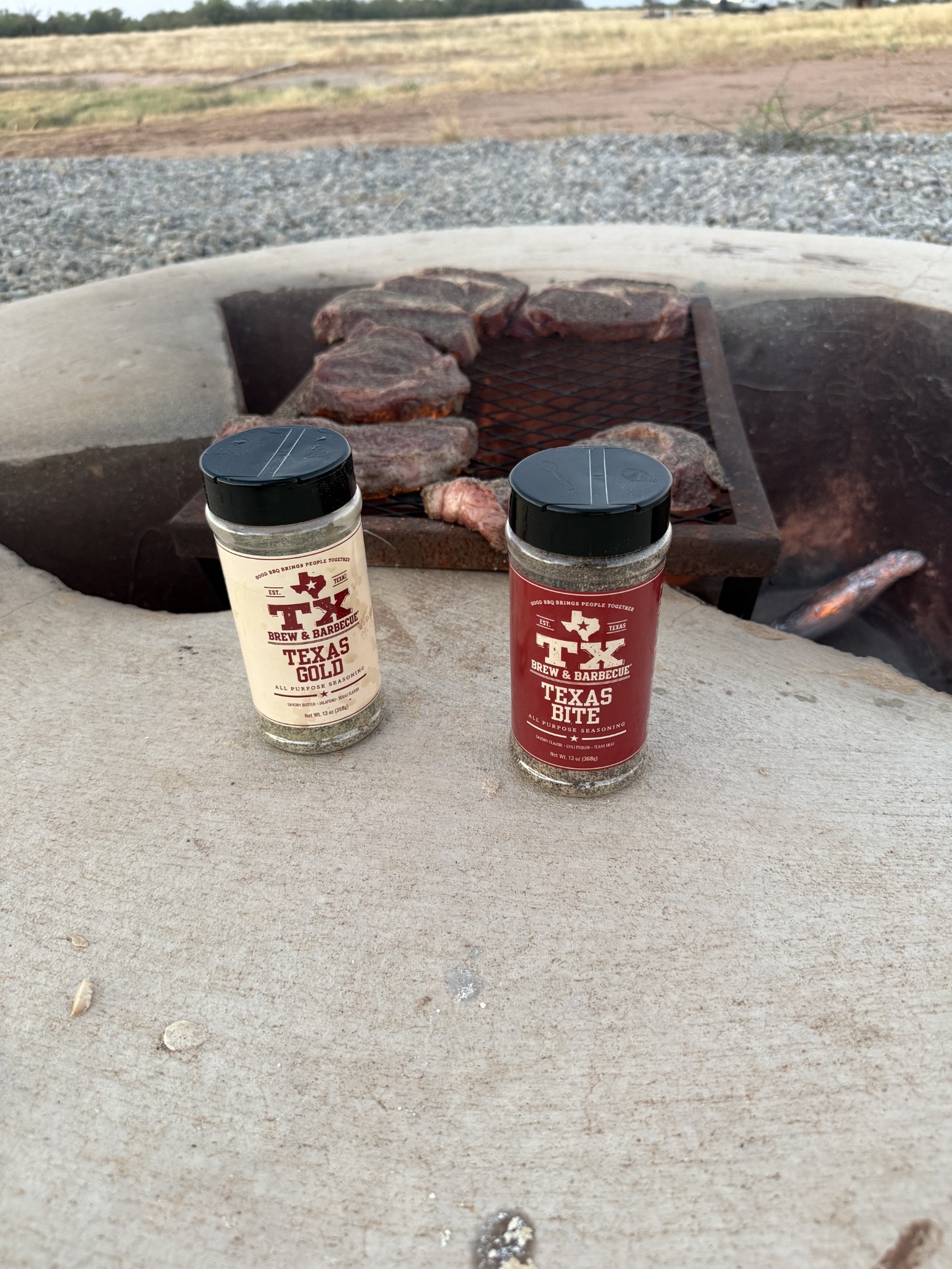 Texas Gold and Texas Bite bottles beside steaks cooking over a fire pit