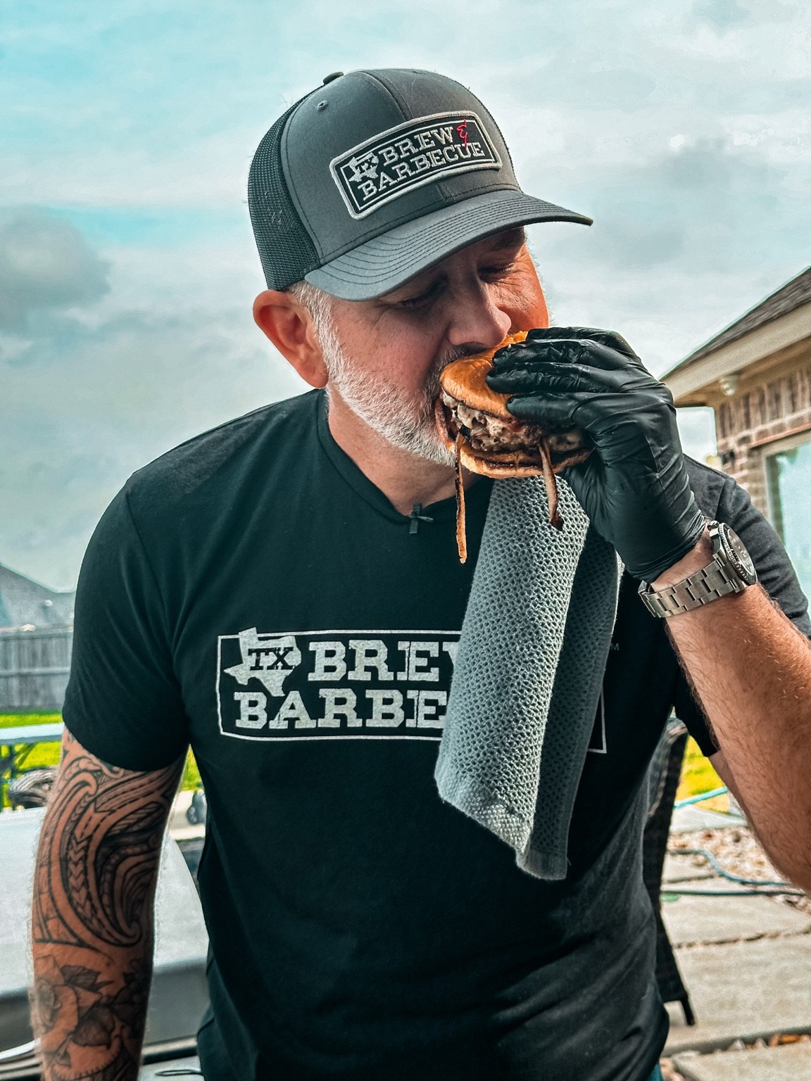 Ty in his TX Brew & Barbecue hat and shirt, taking a bite of a burger.